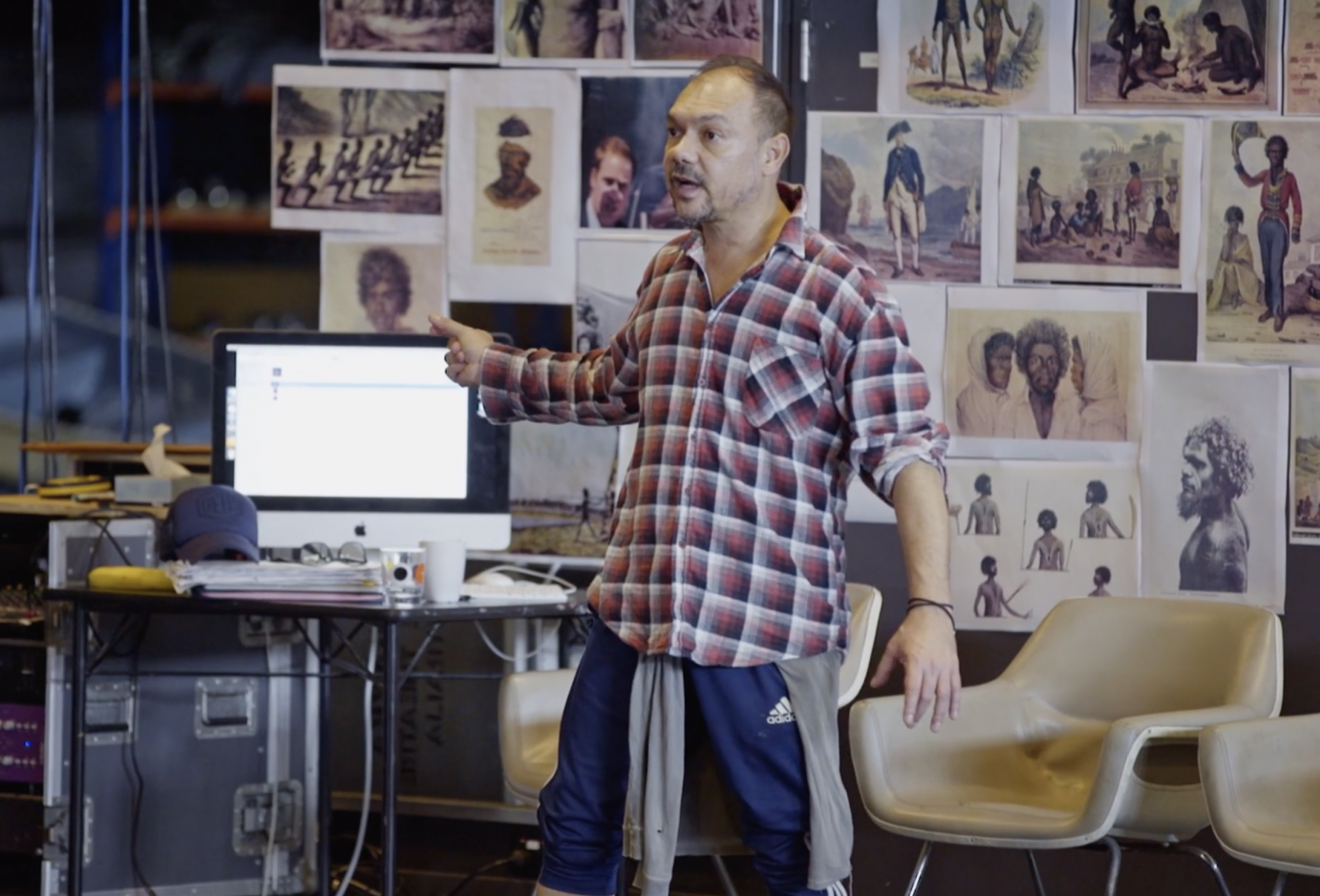 A man in a flannel  shirt stands in front of a wall of pictures, directing dancers